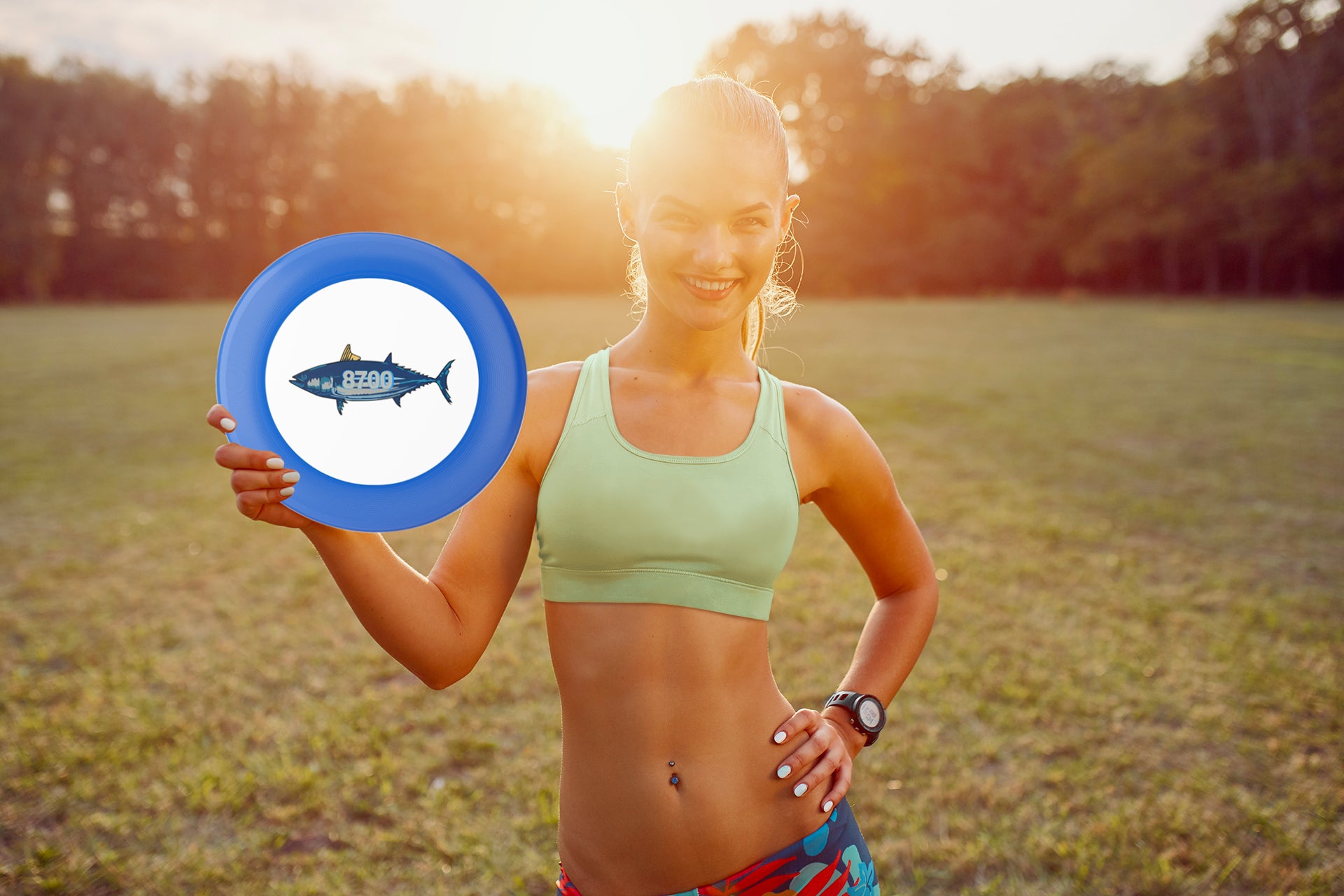 Woman holding a blue and white frisbee with a tuna illustration, smiling in a sunny field